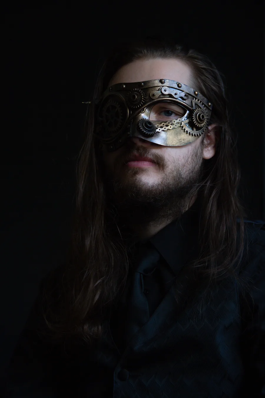 Press photo of Tree of Alchemical Notability: a long-haired man in a black shirt and tie, wearing a brass steampunk mask with gears over one eye